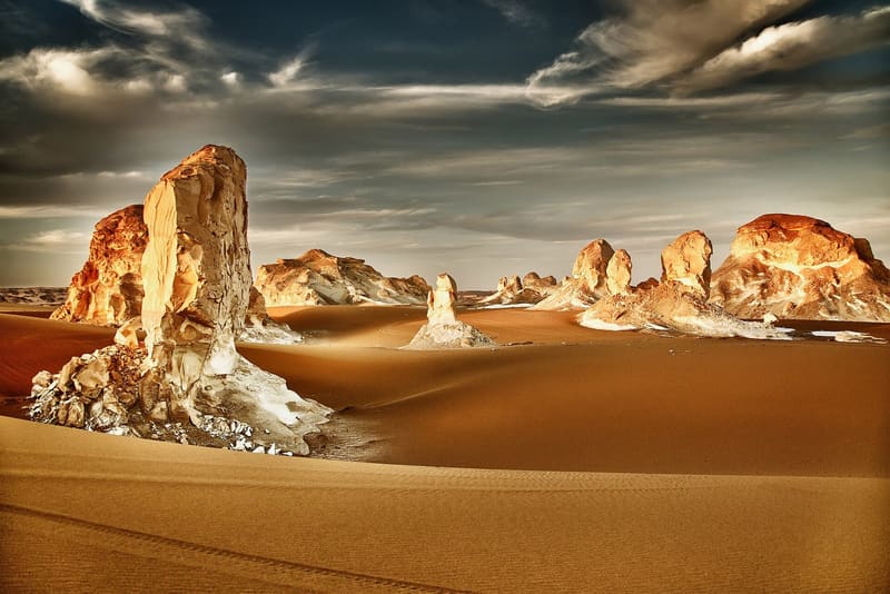 Pinnacoli calcarei illuminati dal sole tra dune dorate sotto nuvole drammatiche